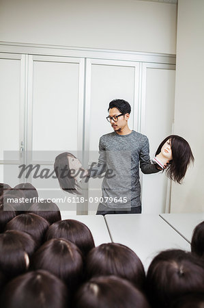 Bearded man wearing glasses standing indoors, holding mannequin head with brown wig in each hand.