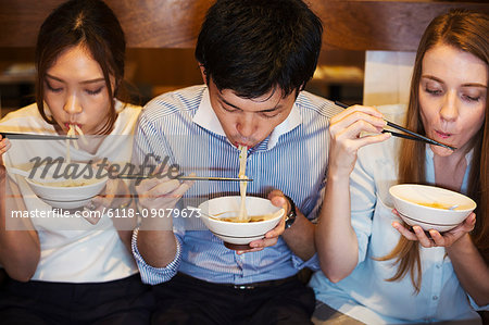 High angle view of three people sitting sidy by side at a table in a restaurant, eating from bowls using chopsticks.