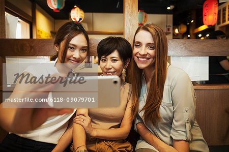 Three women sitting side by side in a restaurant, taking selfie with smartphone.