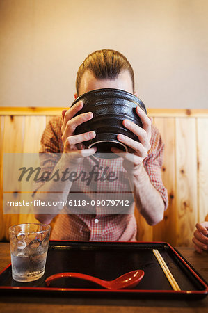 A western man in a noodle restaurant, with a noodle bowl lifting it with two hands to drink from it.