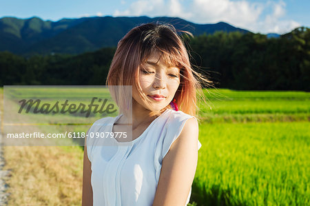 A young woman in a white shirt, hair blown in the wind, standing in open space by rice paddy fields.