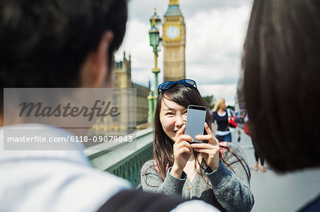 Smiling woman with black hair taking picture of couple with smartphone, standing on Westminster Bridge over the River Thames, London, with the Houses of Parliament and Big Ben in the background.
