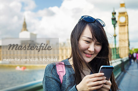 Smiling woman with black hair looking at smartphone, standing on Westminster Bridge over the River Thames, London, with the Houses of Parliament and Big Ben in the background.