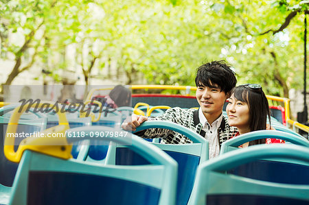 Smiling man and woman with black hair sitting on the top of an open Double-Decker bus driving along tree-lined urban road.