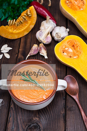 thick pumpkin soup in a ceramic plate on a brown wooden table, behind a fresh pumpkin and garlic, top view