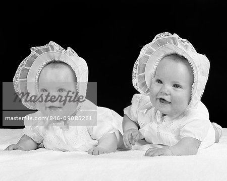 1950s TWIN BABY GIRLS WEARING BONNETS LYING ON THEIR STOMACHS SMILING