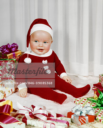 1960s HAPPY BABY WEARING SANTA CLAUS FOOTED PAJAMAS SITTING AMONG WRAPPED CHRISTMAS PRESENTS