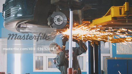 Car auto service - worker grinding metal construction with a circular saw under bottom of the vehicle, telephoto