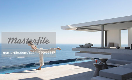 Woman practicing yoga warrior 3 pose on sunny modern, luxury home showcase exterior patio with ocean view