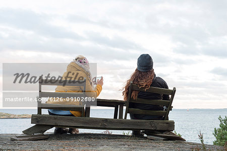 Rear view of teenage girl and young woman sitting on wooden chairs by lake and taking pictures