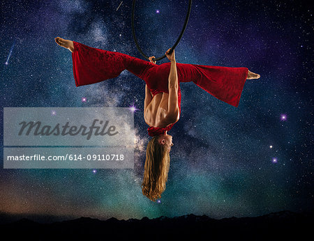 Young female aerial acrobat doing splits hanging upside down from hoop, milky way background