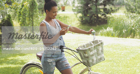 Side view of young woman posing with bicycle on background of green summer park and browsing smartphone cheerfully.