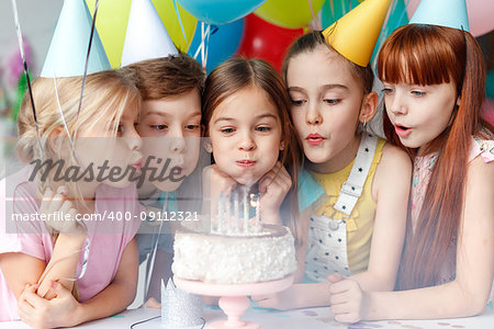 Festive children in party caps, blow candles on delicious cake, make wish, celebrate birthday, have party together, hold colourful balloons. Happy small girl spends festive event with best friends.