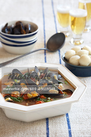 Serving bowl of mussel soup with dinner rolls and beer on a blue striped linen tablecloth