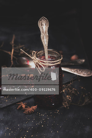 raspberry jam in a glass jar with an iron spoon inside on a black table, behind a toast with jam