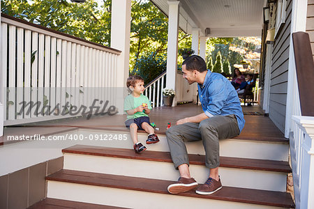 Father And Son Sit On Porch Of House Playing With Toys Together