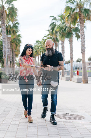 Mature hipster couple strolling while looking at smartphone on sidewalk, Valencia, Spain