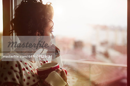 Young woman looking out of window, holding hot drink, thoughtful expression