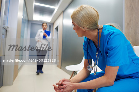 Surgeon sitting in hospital hallway, leaning forward, doctor walking along hallway