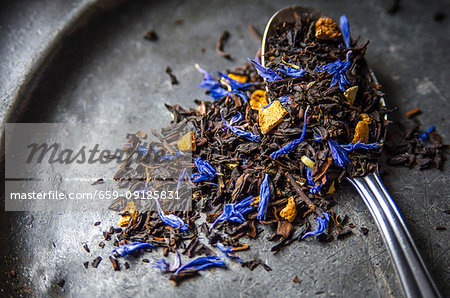 Loose leaf Earl Grey tea with dried citrus peel of lemon, orange and bergamot and blue cornflowers on a dark metal plate and a teaspoon