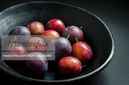 bowl of fresh purple and red prunes and plums in a black fruit bowl on a black background