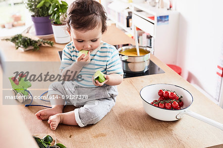 Baby girl sitting on kitchen counter, eating cucumber