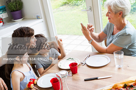 Grandmother sitting at kitchen, photographing grown daughter and grandchildren, using smartphone