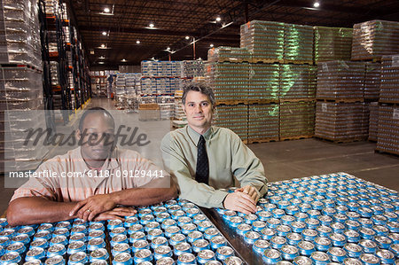 Portrait of an African American male warehouse foreman  and a Caucasian male management person in a warehouse full of cans of flavoured water stored on pallets in the warehouse of a bottling plant.