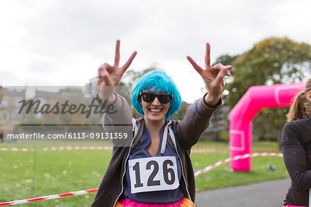 Portrait enthusiastic female runner in wig gesturing peace sign at charity run in park