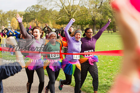 Enthusiastic female runners in tutus crossing charity run finish line in park, celebrating