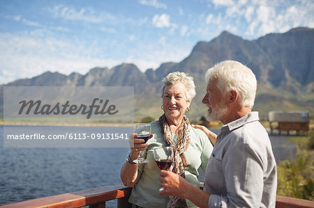 Smiling active senior couple drinking red wine on sunny summer lake balcony