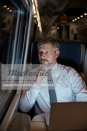Serious, thoughtful businessman working at laptop, looking out window on passenger train at night