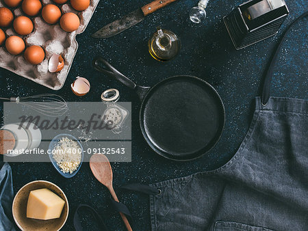 Empty cast-iron frying pan and ingredients for omlette or scrambled eggs,top view.Egg,shell,milk,parmesan cheese,scented rosemary salt,olive oil,pepper,apron,whisk,vintage knife ondark table.Copyspace