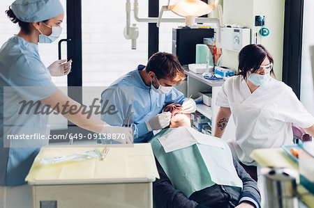 Dentist looking into male patient's mouth, dental nurses preparing equipment