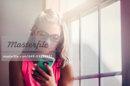 Young girl wearing glasses, beside window, using smartphone