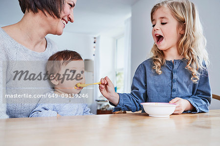 Family sitting at kitchen table, young girl spoon-feeding baby sister