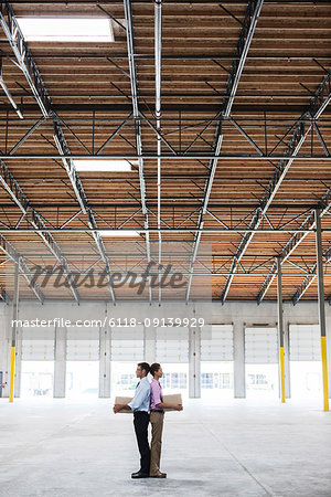 Caucasian man and black female holding cardboard boxes in middle of empty warehouse interior.