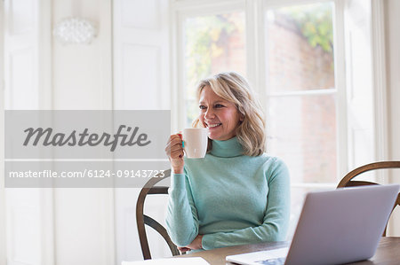 Smiling mature female freelancer drinking tea and working at laptop at home