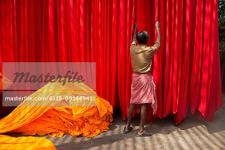 Rear view of man hanging up vibrant and bright red fabric, heap of orange fabric on floor.