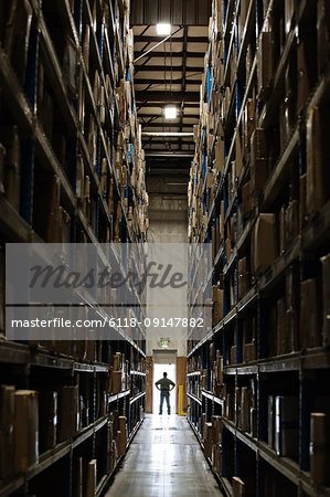 Single warehouse worker in between large racks of cardboard boxes on pallets in a large distribution warehouse;