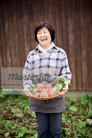 Woman with black hair wearing checkered shirt standing in a garden, holding basket with fresh vegetables, smiling at camera.