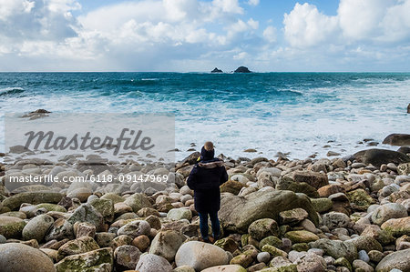 A woman standing on the rocks looking out over the Cornish coastline, and breaking waves.