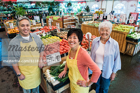 High angle view of man and two women wearing aprons standing at stall with fresh tomatoes at a fruit and vegetable market.