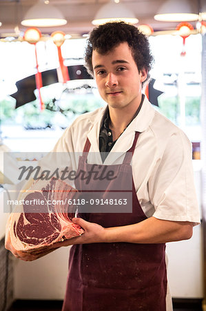Man with brown curly hair wearing apron standing in butcher shop, holding beef forerib, smiling at camera.
