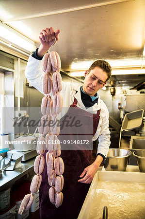 Man wearing apron standing in butcher shop, holding large number of freshly made sausages hanging from hooks.