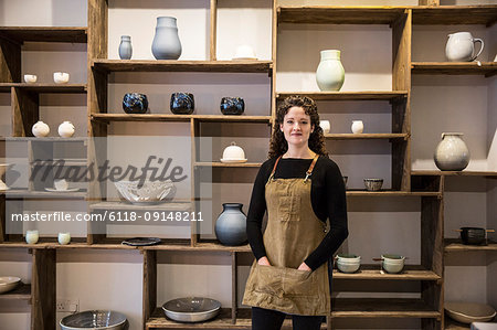 Woman with curly brown hair wearing apron standing in her pottery shop, smiling at camera.