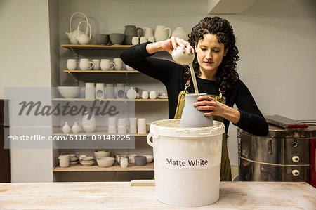 Woman with curly brown hair wearing apron standing in pottery workshop, pouring white glaze over unfired vase.