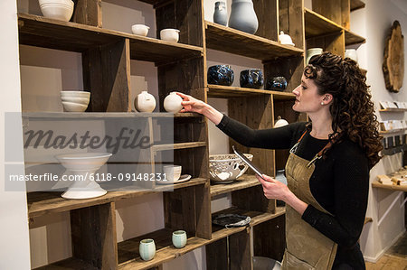 Woman with curly brown hair wearing apron standing in her pottery shop, arranging ceramic items on shelves, holding digital tablet.