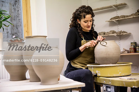 Woman with curly brown hair wearing apron working on spherical clay vase on pottery wheel.