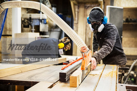 Man wearing ear protectors, protective goggles and dust mask standing in a warehouse, cutting piece of wood with circular saw.